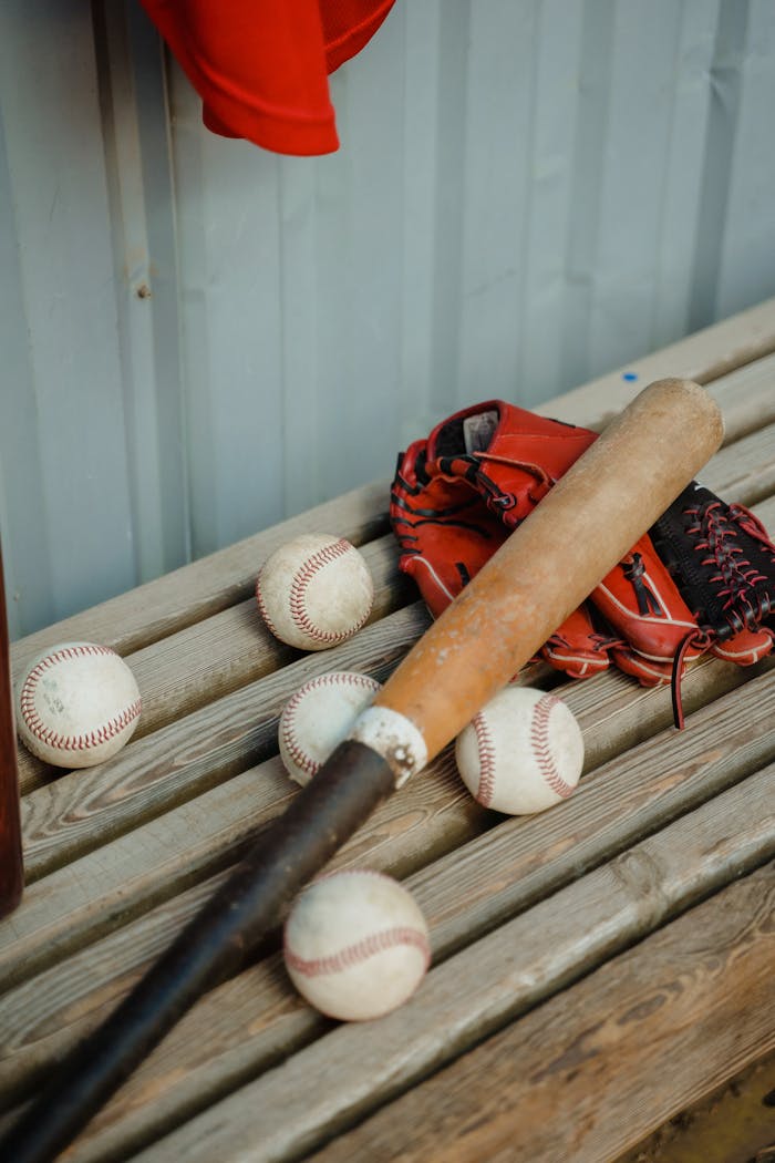 A baseball bat, balls, and a glove on a wooden bench outdoors, showcasing classic sports gear.